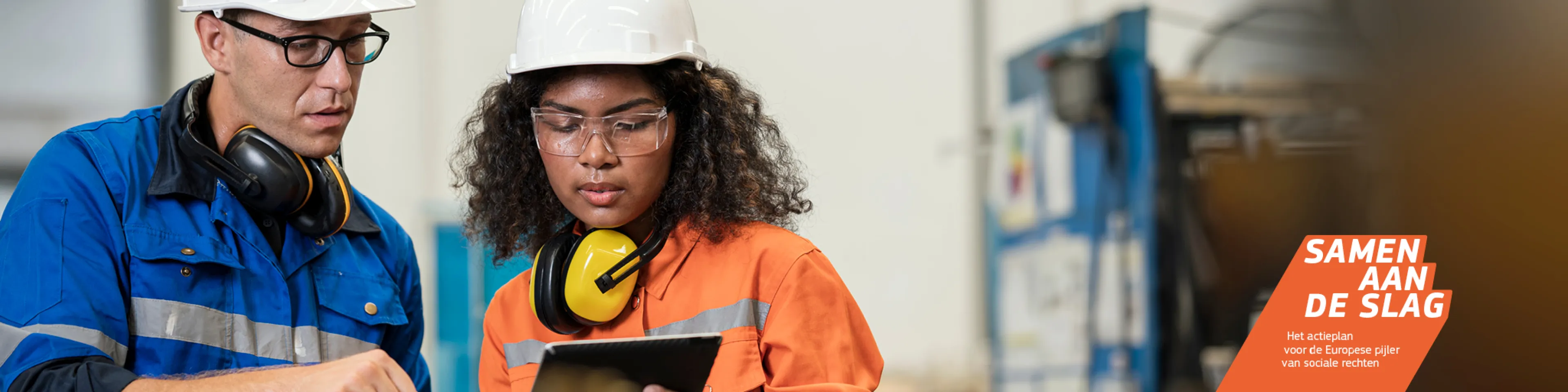 Jonge medewerker met een tablet, in gesprek met een oudere collega in een fabriek. Allebei dragen ze beschermende kleding, met onder meer een veiligheidshelm en -bril.