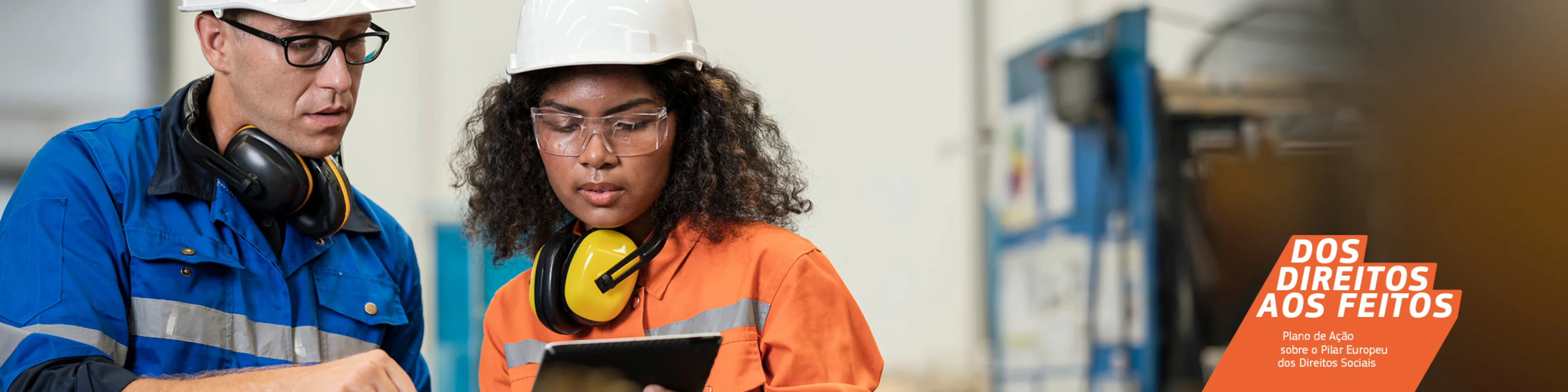 Trabalhador jovem a segurar num tablet e a falar com um trabalhador mais sénior num ambiente de fábrica. Ambos estão com equipamento de proteção, incluindo capacetes e óculos de proteção.