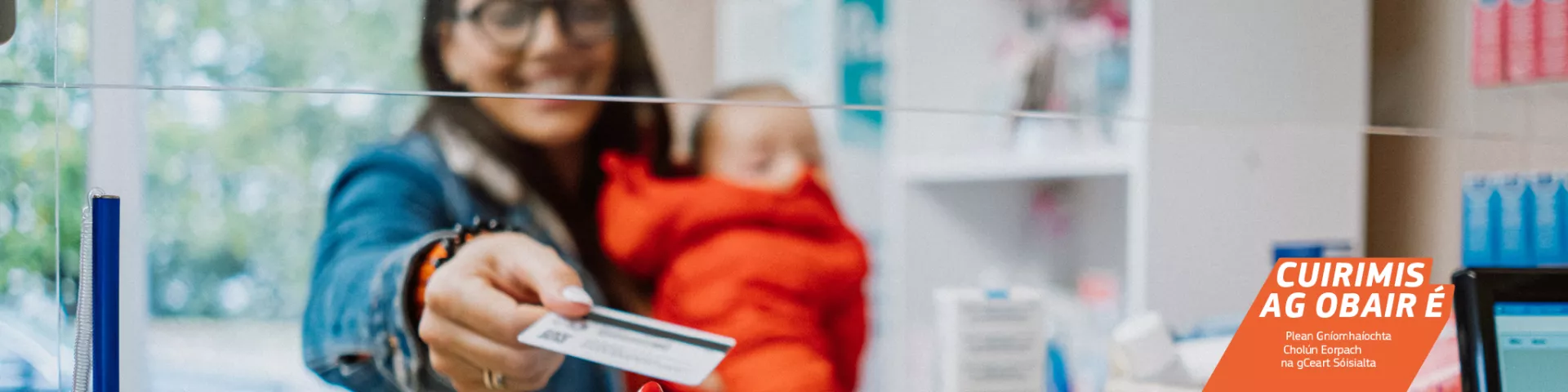 Bean agus babaí ina lámha aici ag déanamh íocaíochta le cárta sa chógaslann