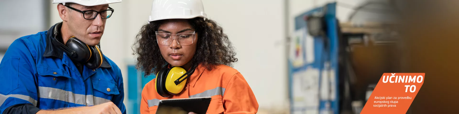 Mlada radnica drži tablet i razgovara s nadređenim radnikom u tvornici. Oboje nose zaštitnu opremu, uključujući kacige i zaštitne naočale.